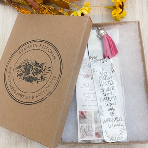 May include: A silver bookmark with a pink tassel, featuring the text "A truly amazing teacher is hard to find, difficult to part with, & impossible to forget." The bookmark is packaged in a brown box with a white interior.