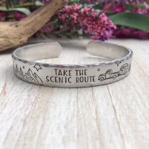 May include: Silver bracelet with the text "Take the Scenic Route" and a small truck pulling a camper. The bracelet has a hammered texture.