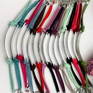 May include: A collection of 14 personalized bracelets with silver metal bands and colorful suede cords. Each band is engraved with a different message. The bracelets are tied with a knot and have a silver clasp.