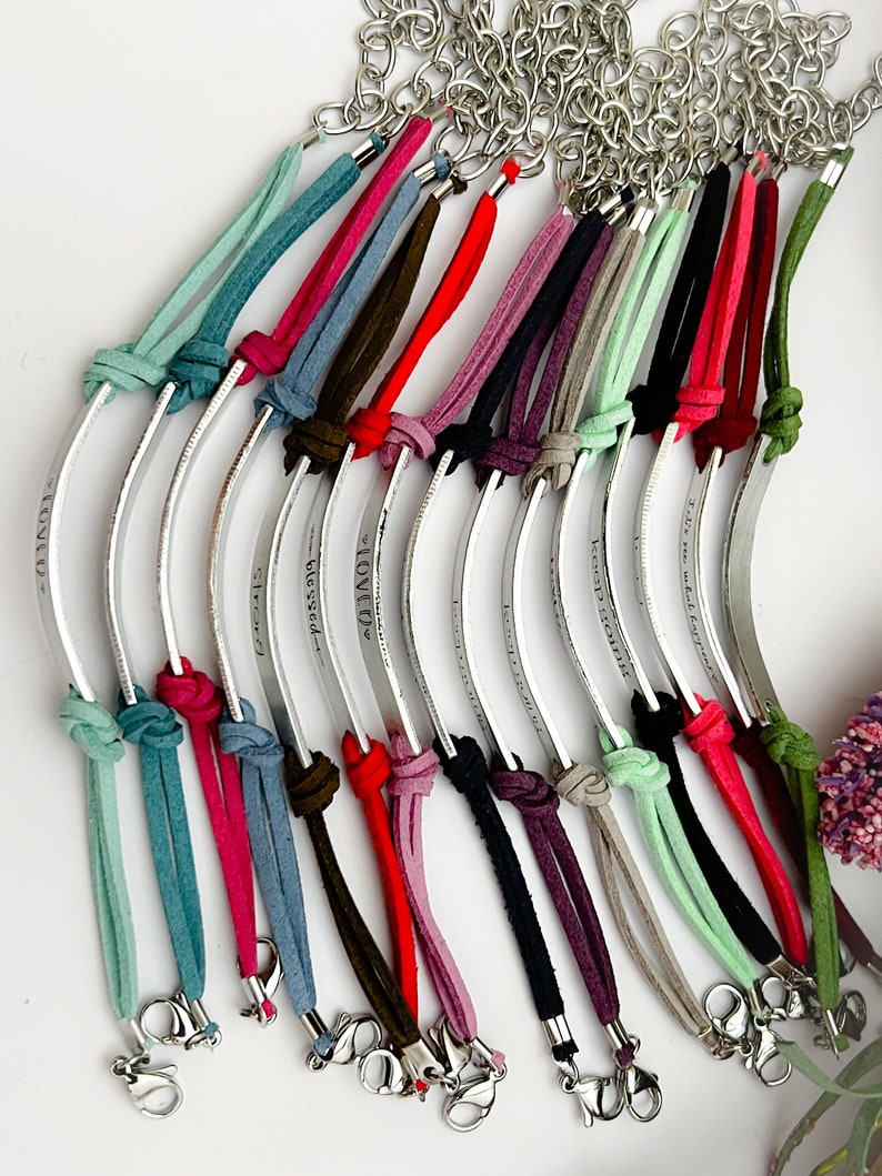 May include: A collection of 14 personalized bracelets with silver metal bands and colorful suede cords. Each band is engraved with a different message. The bracelets are tied with a knot and have a silver clasp.