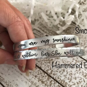 May include: Two silver bracelets with different finishes. The top bracelet has a smooth finish and the text "you are my sunshine" engraved on it. The bottom bracelet has a hammered edge and the text "is within her. She will not f" engraved on it.