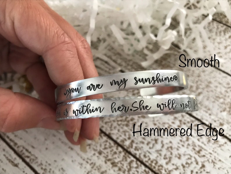 May include: Two silver bracelets with different finishes. The top bracelet has a smooth finish and the text "you are my sunshine" engraved on it. The bottom bracelet has a hammered edge and the text "is within her. She will not f" engraved on it.
