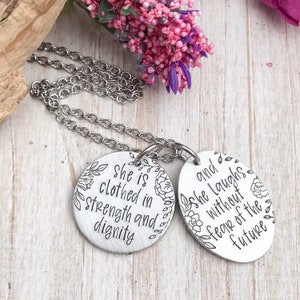 Peut inclure: Un collier en argent avec deux pendentifs ronds. Le pendentif de gauche porte le texte "She is clothed in strength and dignity" avec des motifs floraux. Le pendentif de droite porte le texte "and she laughs without fear of the future" avec des motifs floraux.