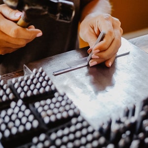 Op de afbeelding: Een persoon gebruikt een metalen gereedschap om een metalen staaf te markeren. De staaf ligt op een metalen oppervlak. Op de achtergrond zijn meerdere trays met kleine metalen stempels te zien.