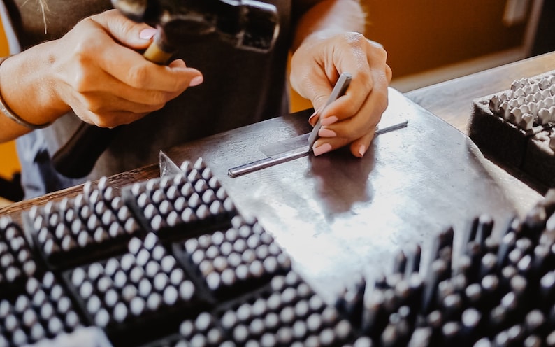 Op de afbeelding: Een persoon gebruikt een metalen gereedschap om een metalen staaf te markeren. De staaf ligt op een metalen oppervlak. Op de achtergrond zijn meerdere trays met kleine metalen stempels te zien.