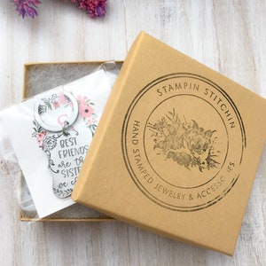 May include: A silver keychain with a floral design and the text "Best Friends are the Sisters we choose". The keychain is packaged in a brown box with a black and white logo that says "Stampin Stitch'n Hand Stamped Jewelry & Accessories".