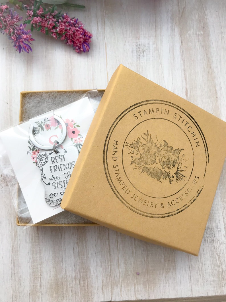 May include: A silver keychain with a floral design and the text "Best Friends are the Sisters we choose". The keychain is packaged in a brown box with a black and white logo that says "Stampin Stitch'n Hand Stamped Jewelry & Accessories".