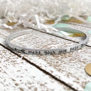 May include: Silver bangle bracelet with the inscription "make your own path" and an arrow pointing to the right.