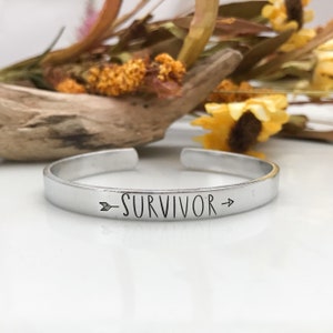 May include: A silver metal bracelet with the word "SURVIVOR" engraved on it, with arrows pointing to the left and right of the word.