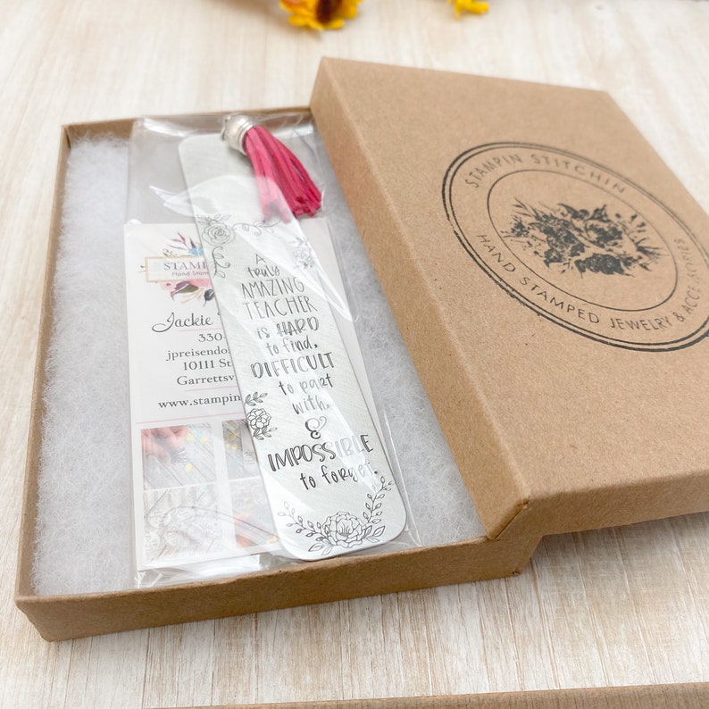 May include: A silver bookmark with a pink tassel, packaged in a brown box. The bookmark has a quote that reads "A truly amazing teacher is hard to find. Difficult to part with. & Impossible to forget."