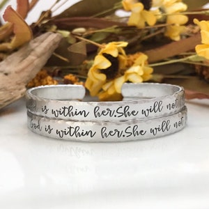 May include: Two silver bracelets with the inscription "God is within her. She will not" engraved on them. The bracelets are stacked on top of each other.