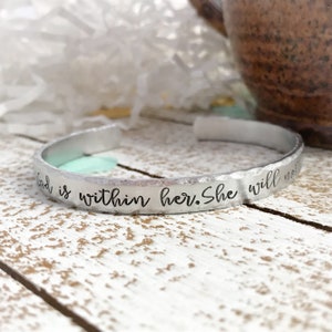 May include: A silver metal bracelet with the words "God is within her. She will not"
