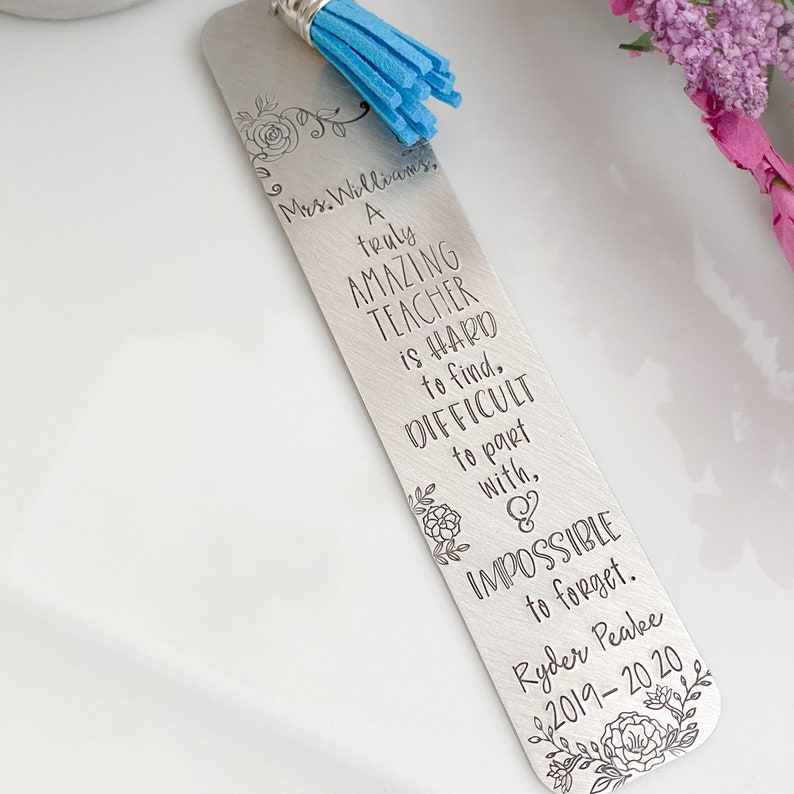 Op de afbeelding: Zilveren bladwijzer met een blauwe kwast en een bloemmotief. De bladwijzer heeft een gegraveerd bericht dat luidt: "Mrs. Williams, een echt GEWELDIGE LERAAR is MOEILIJK te vinden, MOEILIJK om afscheid van te nemen, en ONMOGELIJK om te vergeten. Ryder Peake 2019-2020"