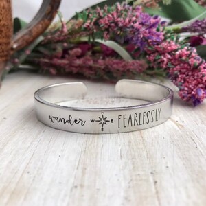 May include: A silver metal cuff bracelet with the words "wander fearlessly" and a compass rose engraved on it.