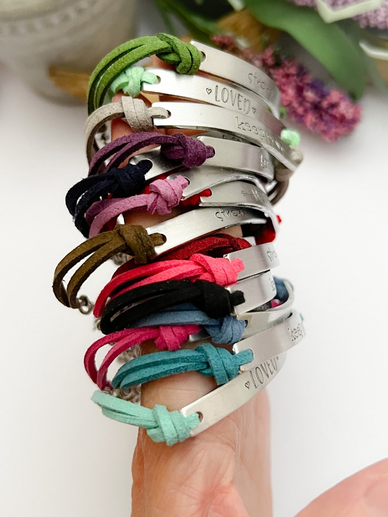 May include: A stack of 12 leather bracelets with silver metal charms. Each charm has a different engraved message, including "Loved", "Strong", and "Keep Going".