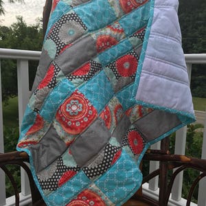 May include: A handmade quilt with a patchwork design. The quilt features squares of teal, gray, red, and black fabrics with floral and polka dot patterns. The quilt is trimmed with a teal border and white backing.