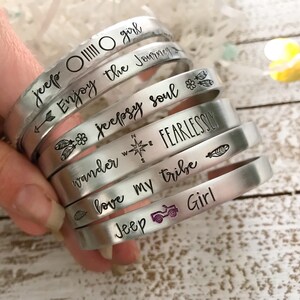 May include: A set of silver metal bracelets with various engraved sayings, including "jeep OIIIII O girl", "Enjoy the Journey", "jeepsy soul", "wander FEARLESSLY", "love my tribe", and "Jeep Girl".