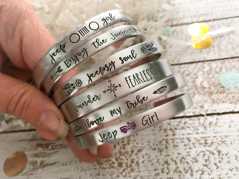 May include: A set of silver metal bracelets with various engraved sayings, including "jeep OIIIII O girl", "Enjoy the Journey", "jeepsy soul", "wander FEARLESSLY", "love my tribe", and "Jeep Girl".