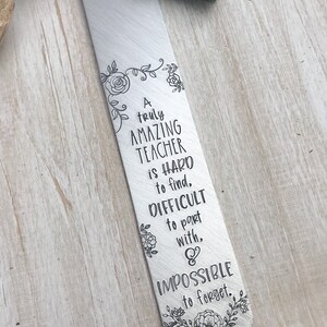 May include: A silver bookmark with a teal tassel. The bookmark features a floral design and the text "A truly AMAZING TEACHER is HARD to find, DIFFICULT to part with, & IMPOSSIBLE to forget."