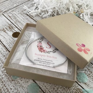 May include: A silver bangle bracelet in a brown cardboard box with a white floral design and the text "Stampin Stitchn" and contact information for Jackie Preiendofer.