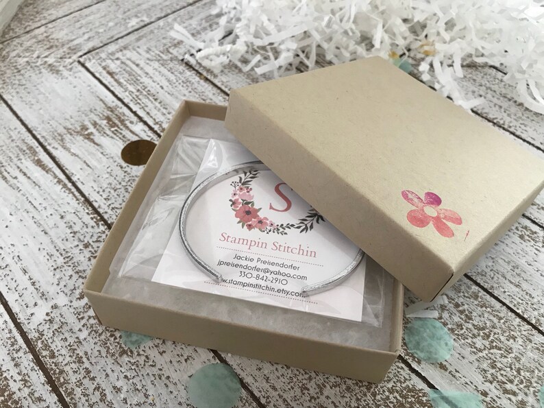May include: A silver bangle bracelet in a brown cardboard box with a white floral design and the text "Stampin Stitchn" and contact information for Jackie Preiendofer.
