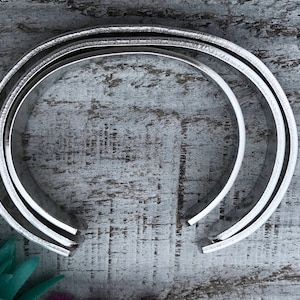 May include: Two silver metal bracelets stacked on top of each other. The bracelets are round and have a smooth, polished finish.