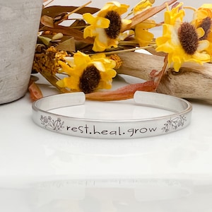 Pode incluir: Uma pulseira de punho prateada com as palavras "rest.heal.grow" gravadas. A pulseira apresenta desenhos florais em cada extremidade. A pulseira é exibida com girassóis amarelos e folhagem seca. A pulseira é de metal.