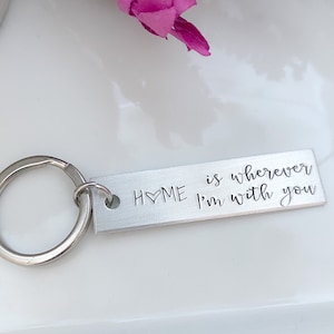 May include: A silver rectangular keychain with the words "HOME is wherever I'm with you" engraved on it. The keychain has a silver ring attached. Pink and purple flowers are in the background.