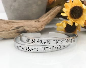 May include: Two silver bracelets with engraved coordinates. The top bracelet reads "41° 24' 40.6"N 75° 37' 44.8°W" and the bottom bracelet reads "41° 25' 23.6"N 75° 37' 22.1 W".