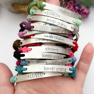 May include: A stack of bracelets with different colored cords and silver charms. The charms have inspirational messages engraved on them, including "loved", "keep going", "strong", and "blessed".