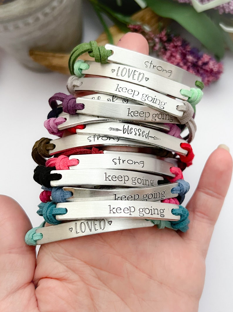 May include: A stack of bracelets with different colored cords and silver charms. The charms have inspirational messages engraved on them, including "loved", "keep going", "strong", and "blessed".