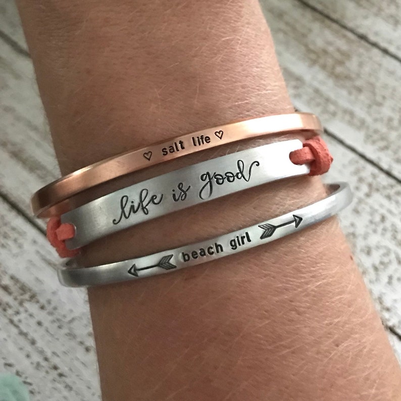 May include: Three silver bracelets with engraved text. The top bracelet reads "salt life" with a heart symbol on each side. The middle bracelet reads "life is good" in cursive. The bottom bracelet reads "beach girl" with an arrow pointing to the right.
