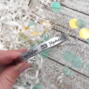 May include: Silver bracelet with the words "Capture Moments" and a camera icon engraved on it.