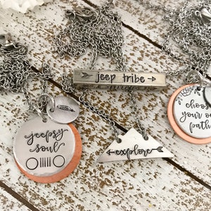 May include: Three silver necklaces with different charms. The first necklace has a bar charm that says "jeep tribe" with arrows on either side. The second necklace has a round charm that says "jeeps soul" with a jeep grill design. The third necklace has a round charm that says "choose your own path" with a small arrow charm. All of the charms are on a copper background.