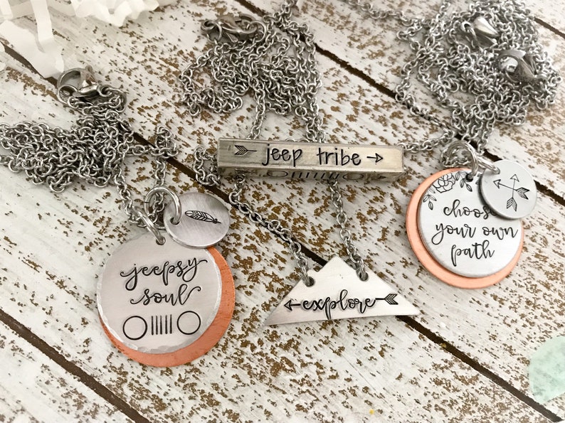 May include: Three silver necklaces with different charms. The first necklace has a bar charm that says "jeep tribe" with arrows on either side. The second necklace has a round charm that says "jeeps soul" with a jeep grill design. The third necklace has a round charm that says "choose your own path" with a small arrow charm. All of the charms are on a copper background.