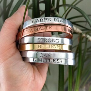 May include: A stack of six silver, copper, and gold bracelets with inspirational messages engraved on them. The messages include "Carpe Diem", "Namaste", "Strong", "Believe", "I Can", and "Limitless".