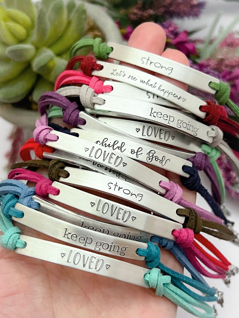 May include: A stack of colorful bracelets with silver metal tags. The tags have inspirational messages engraved on them, including "strong", "keep going", "child of God", and "loved".