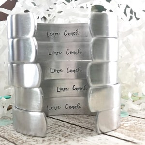 May include: Five silver metal bracelets stacked on top of each other. Each bracelet has the words "Love Coach" engraved on it.