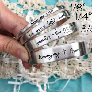May include: Four silver metal bracelets with different engraved sayings. The bracelets are stacked on top of each other and are 1/8", 1/4", 3/8", and 1/2" wide.