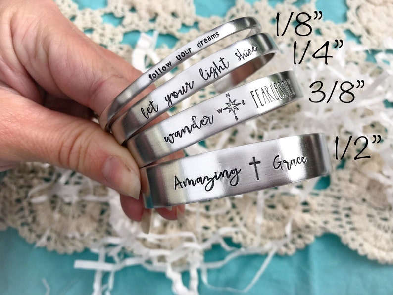May include: Four silver metal bracelets with different engraved sayings. The bracelets are stacked on top of each other and are 1/8", 1/4", 3/8", and 1/2" wide.
