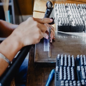 May include: A person uses a metal tool to stamp a metal bar. The bar is resting on a metal surface. There are multiple trays of metal stamps in the background.