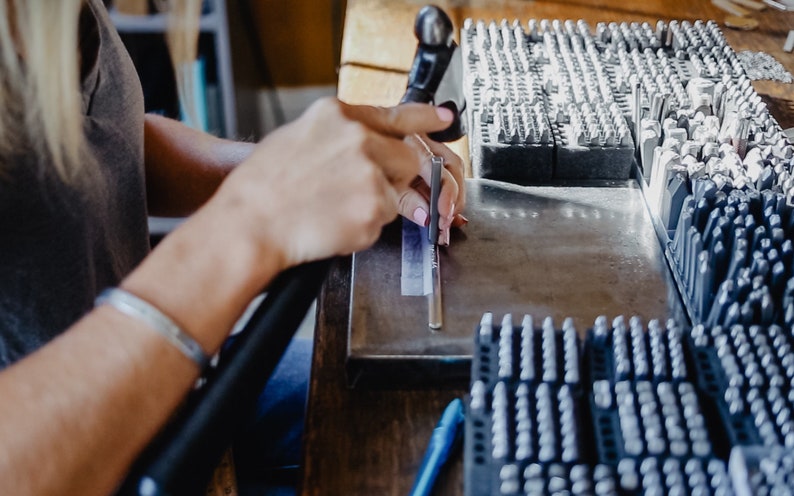 May include: A person is using a metal tool to stamp a metal bar. The bar is resting on a metal surface. There are multiple trays of metal stamps in the background.