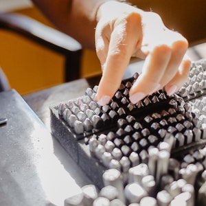 May include: A close-up of a metal stamp set with a variety of different sized and shaped stamps. The stamps are arranged in rows on a black foam base. A hand is reaching down to touch the stamps.