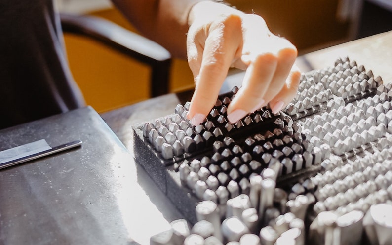 May include: A close-up of a metal stamp set with a variety of different sized and shaped stamps. The stamps are arranged in rows on a black foam base. A hand is reaching down to touch the stamps.