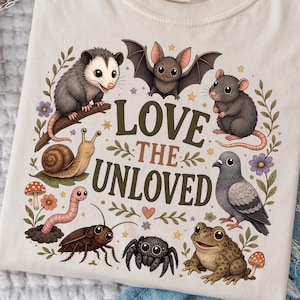 May include: A cream-colored t-shirt featuring a graphic print of an opossum, bat, rat, snail, worm, cockroach, spider, pigeon, and frog surrounding the text LOVE THE UNLOVED in dark green and brown letters, with small floral and star accents.