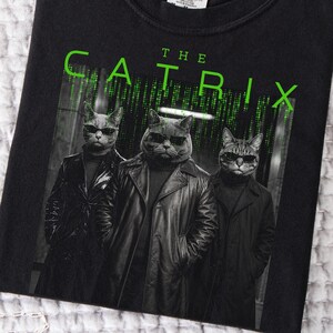 May include: A black t-shirt featuring a graphic of three cats wearing black leather trench coats and sunglasses. Above the cats, the text reads THE CATRIX in bright green letters, set against a background of falling green digital code.
