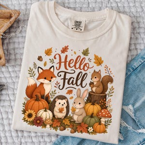 May include: A cream-colored t-shirt featuring a graphic print with the text Hello Fall. The design includes a fox, a squirrel on a tree stump, a hedgehog, and a rabbit surrounded by pumpkins, mushrooms, acorns, and autumn leaves in orange and brown.