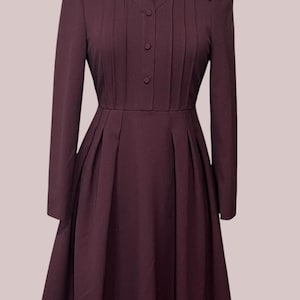 May include: A long-sleeved, dark burgundy dress featuring a pointed collar, a button-down front placket, and vertical pleats extending from the shoulders to the waist. The skirt is pleated at the waist, creating a full, flared silhouette.