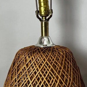 May include: A table lamp featuring a rounded, woven rattan base with a crisscross pattern. The lamp has a brass-colored metal base, a silver-toned neck, and a brass-finished harp holding a single white light bulb at the top of the fixture.