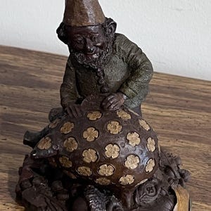 May include: A brown resin figurine of a gnome wearing a pointed hat, leaning over a pig decorated with light-colored flower patterns. The base features sculpted leaves and acorns with the text "© 1984 Clark" and "Tim Clark" inscribed on it.